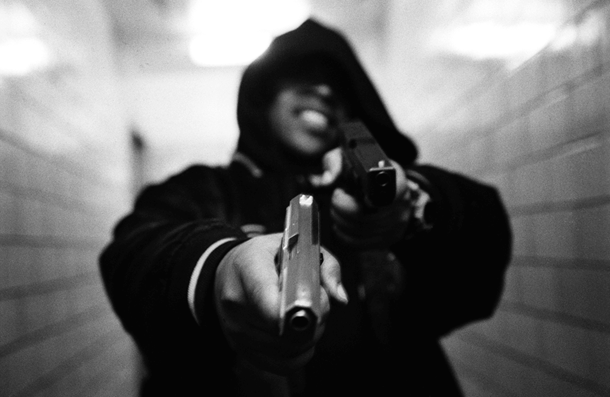 Black-and-white photograph of Boogie, a hooded figure pointing two guns at the camera