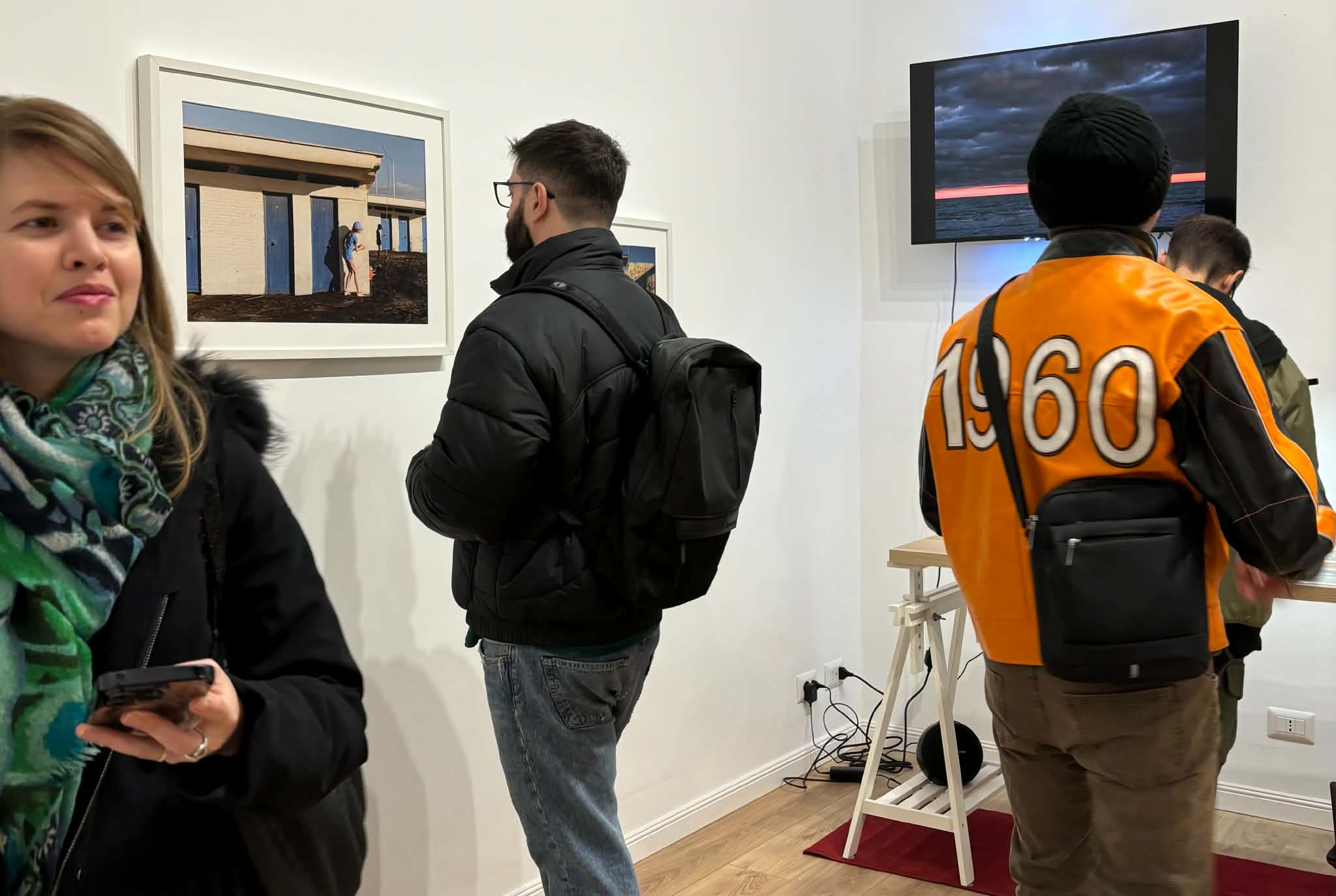 The Daylight School's indoor exhibition hall. A visitor is looking at a framed photograph of beach cabanas, while another visitor, wearing an orange jacket, is watching a video on a monitor. Various artworks and people are in the room.
